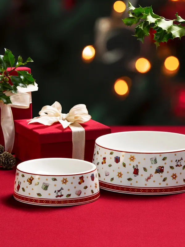 Due ciotole Toy's Delight per animali con motivi natalizi su un tessuto rosso, accanto a due scatole regalo rosse con nastri bianchi e decorazioni verdi.