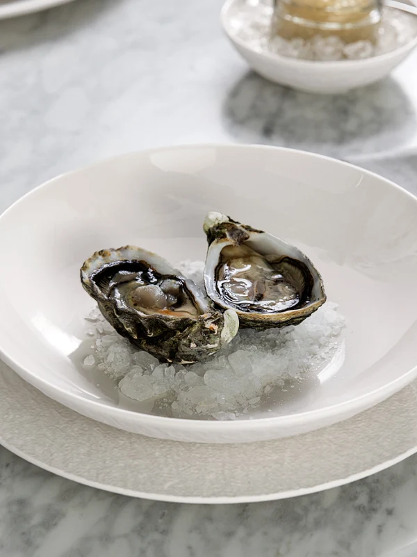 Dos ostras sobre una cama de hielo se sirven en un plato blanco Manufacture, al lado de ellas, un tenedor y una servilleta reposan sobre una mesa de mármol.