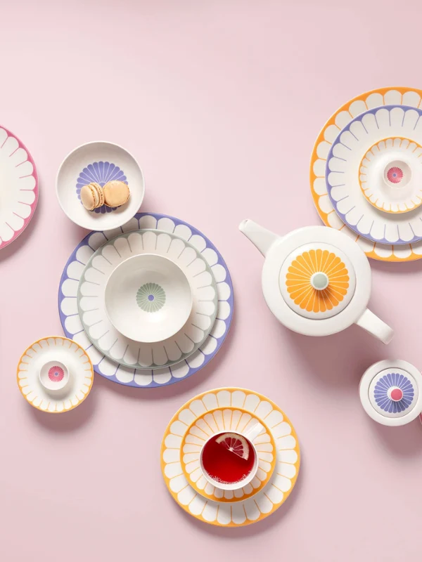 Complete tea set consisting of cups, bowls, and plates with floral patterns from Fleur Various patterned ceramic bowls, dishes, and a teapot from the Fleur collection on a pink background, several plates with colorful desserts.