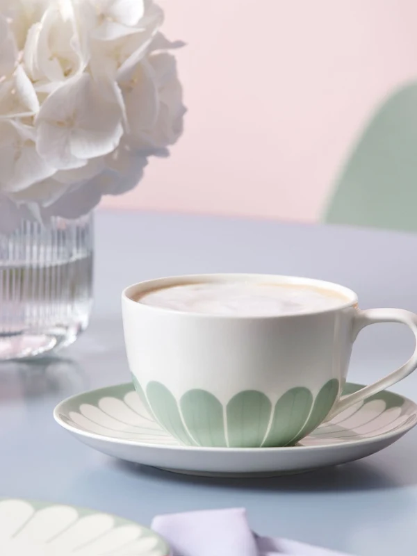 Fleur cup with pastel green floral pattern on matching saucer A white cup with a pastel green leaf pattern from the Fleur collection stands on a matching saucer next to a glass vase with white flowers on a pastel-colored table.