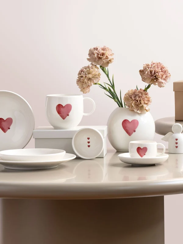 With love Artykuły ceramiczne z motywami serc Naczynia ceramiczne marki with love z czerwonymi motywami serc, ułożone na okrągłych stołach z kwiatami i pudełkiem prezentowym.