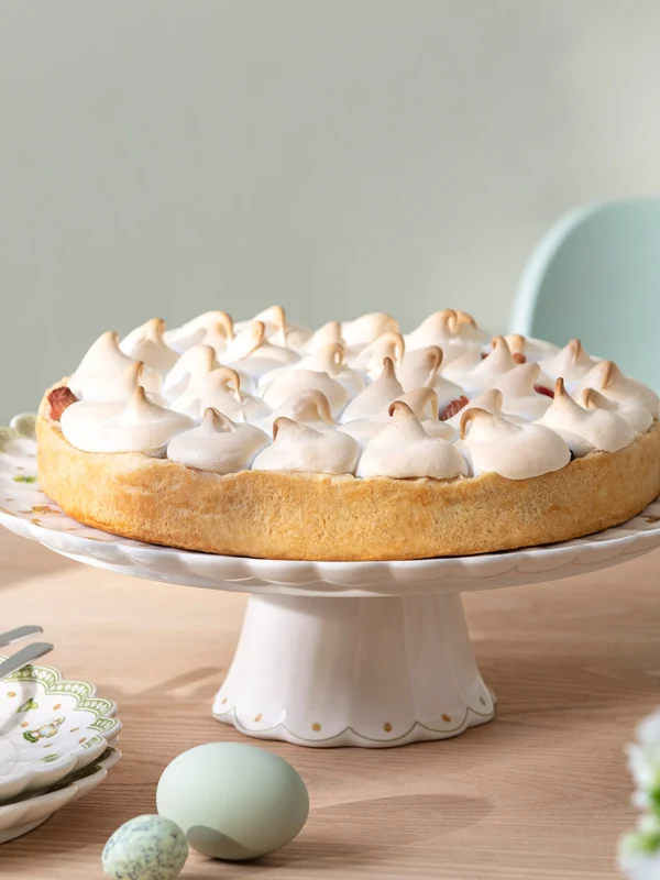 Easter Delight cake stand with Easter decorations A cake with meringue stands on a wooden table on a cake stand with Easter decorations from Easter Delight, next to plates and flowers.