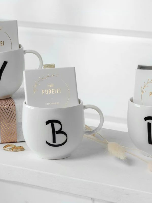 Kubek Letters z literami V, B i P wypełniony biżuterią na półce Trzy spersonalizowane kubki Letters z inicjałami „v”, „b” i „p” wypełnione biżuterią i pudełka z kosmetykami na białej półce.