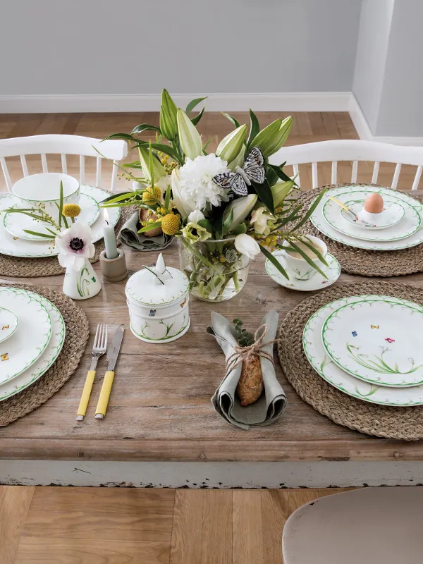 A “Colourful Spring” dinner set for four from Villeroy & Boch consists of porcelain with a floral pattern, flatware with yellow handles, woven placemats, and a floral arrangement. Each place setting includes an egg in a decorative cup.