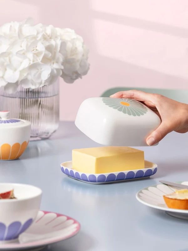 Fleur butter dish with other tableware items from Fleur on a blue table A hand lifts the lid of the Fleur butter dish, which is standing on a table with plates, cups, and a vase from the Fleur collection with white flowers.