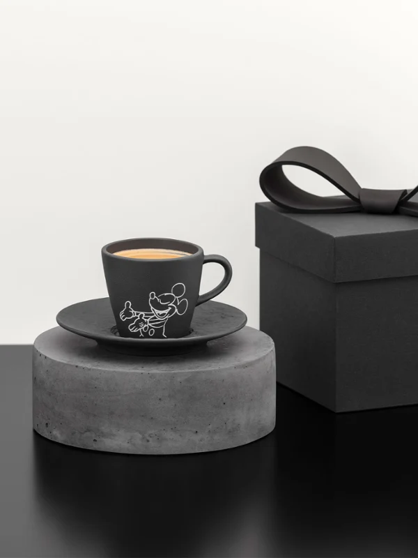 Taza de café negra y platillo sobre un pedestal gris Taza de café negra y platillo sobre un pedestal gris junto a una caja de regalo negra con un lazo sobre una superficie negra.