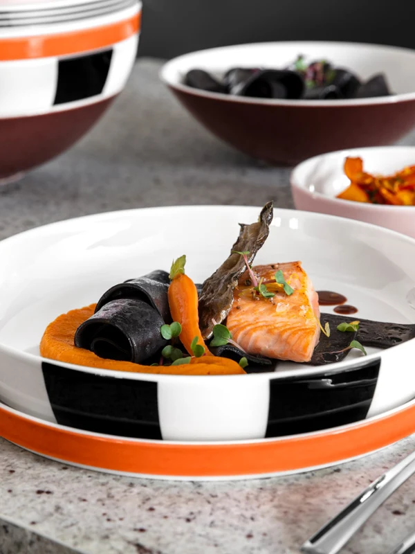 A Memphis-style dish with salmon, carrots, and black noodles, with sides and cutlery on a stone tabletop.