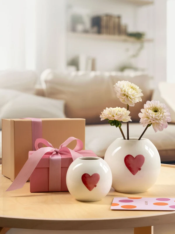 Due vasi bianchi con motivi a cuori rossi di with love, una confezione regalo rosa e un biglietto su un tavolino rotondo in un accogliente salotto.