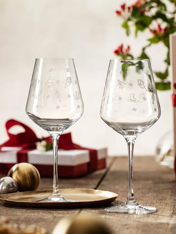 Two Villeroy & Boch Toy's Delight glasses on a wooden table with Christmas presents, a bottle of wine, a pine cone and festive decorations in the background.