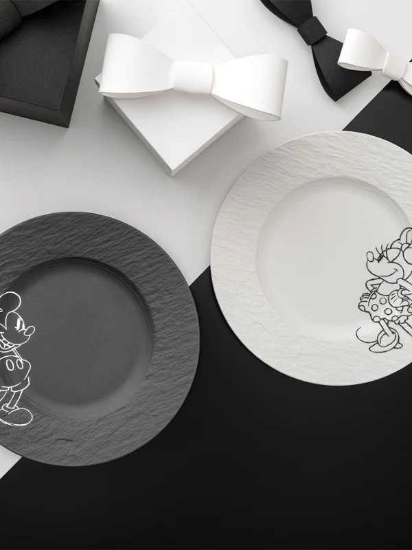Dwa talerze z kolekcji Manufacture Mickey, czarny i biały, stoją na podzielonym czarno-białym tle z dwoma pudełkami prezentowymi z kokardką.
