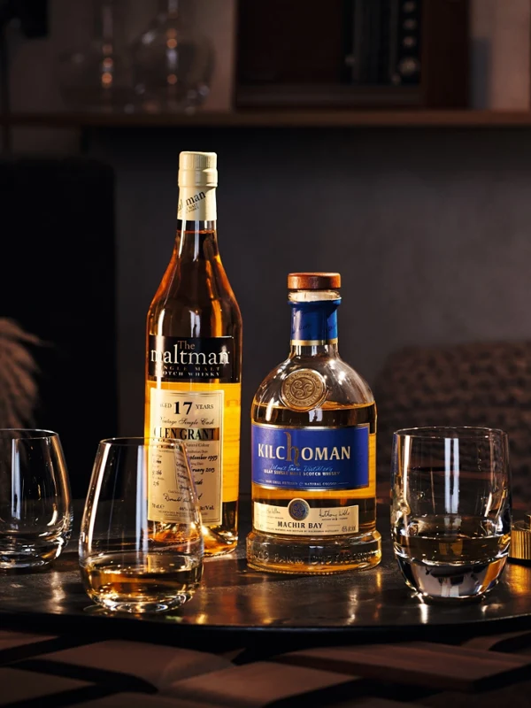 Twee flessen Schotse whisky en drie glazen van Villeroy & Boch staan op een houten tafel. Op de achtergrond zijn boeken en een decoratiefiguur te zien.