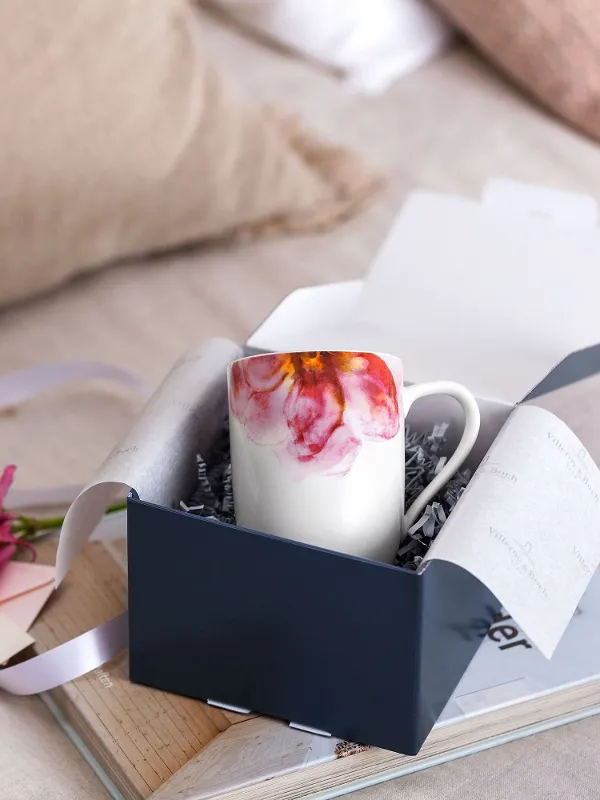 An open Villeroy & Boch gift box with a Rose Garden cup, pink flowers and a note on a wooden tray, with soft cushions in the background.