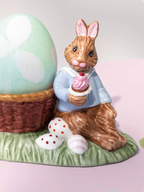 Ceramic Annual Easter Edition 2026 bunny figurine in a blue jacket sits beside a large pastel egg in a basket on a grassy base with small eggs.