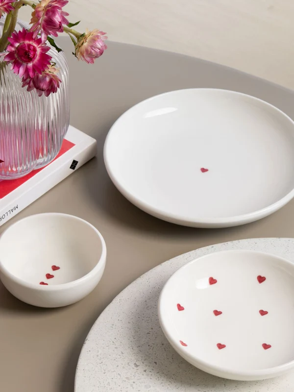 With love Ceramiczne talerze i miseczki z czerwonymi motywami serc Trzy białe ceramiczne talerze z małymi czerwonymi motywami serc z kolekcji with love stoją na stole obok wazonu z różowymi kwiatami i pudełka prezentowego.