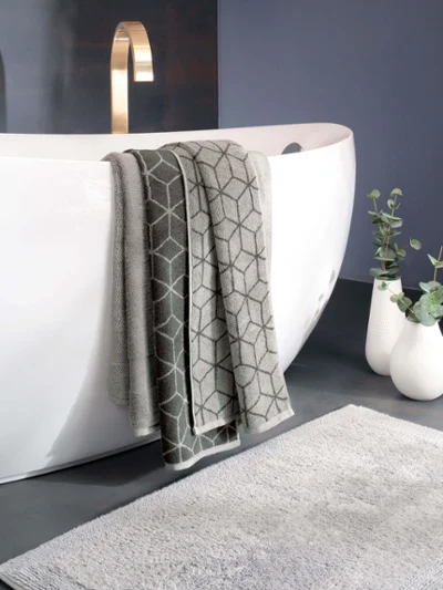 Ręczniki Carré nad białą wanną: Ręczniki Carré nad białą wanną Trzy szare ręczniki wiszą nad białą wanną wolnostojącą ze złotą baterią. Na podłodze leży szara mata łazienkowa, obok stoją dwa białe wazony z zielonymi roślinami.