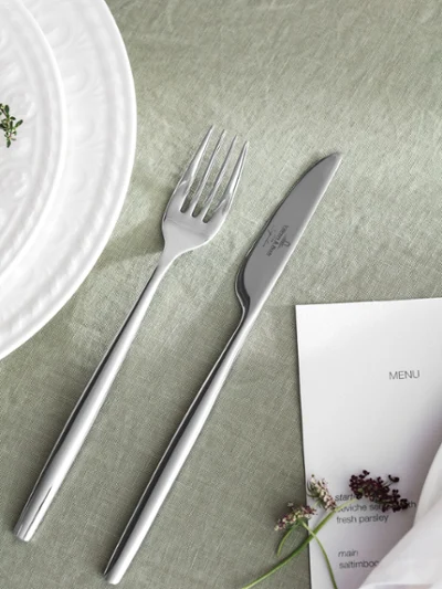 Stainless steel dinner fork and knife arranged beside a white embossed porcelain dinner plate on a soft green linen tablecloth, with a printed menu and fresh herb sprigs creating an elegant place setting.