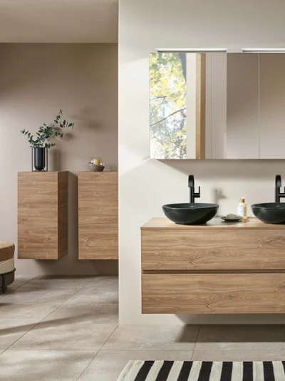Un plan de toilette Arto au look bois et un lavabo noir Une salle de bains moderne avec des placards en bois, un double lavabo, un grand miroir et une fenêtre avec vue sur les arbres.