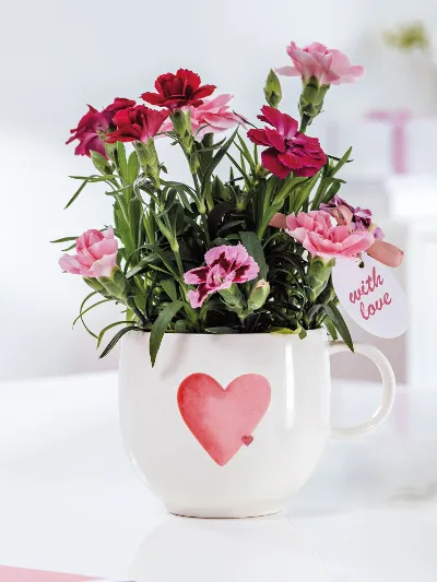 A white With Love cup with a red heart and pink and red flowers on a white table.