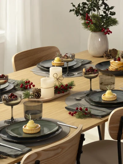 Lave Table set for a meal with plates, cutlery, candles, wine glasses, and a vase with greenery and red berries.