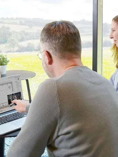 Miejsce pracy z dwiema osobami Dwie osoby siedzą przed laptopem. Mężczyzna wskazuje na ekran.