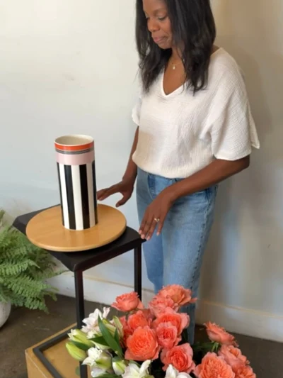 The start of a floral arrangement with Memphis vase Woman arranging a vibrant bouquet of coral roses and white lilies beside a modern striped vase, creating a warm, elegant home setting.