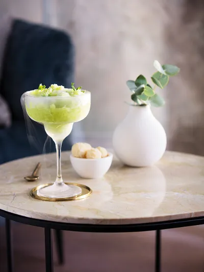 A chilled cocktail garnished with lime zest, served in a Purismo cocktail glass on a marble table, accompanied by a small bowl of snacks and a white vase with green plants.