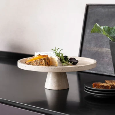 Plat de service parfait en travertin pour antipasti ou fromage. Un assortiment de fromages sur un plat de service en travertin avec pied, et des fruits foncés, à côté d'une vaisselle noire Manufacture de Villeroy & Boch sur un plan de travail foncé.