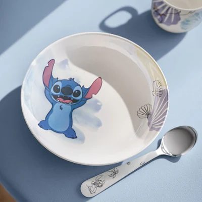 Eine weiße Disney Stitch Schale, ein passender Löffel und eine Tasse auf einem Tisch Eine weiße Disney Stitch Schale mit einem Cartoon-Motiv, ein passender Löffel und eine Tasse auf einem blauen Tisch im Sonnenlicht.