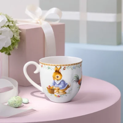 A decorative Annual Easter Edition 2026 mug with a bunny design sits on a pink table next to a gift box and a vase of white flowers.