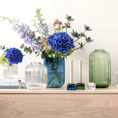 A sideboard with Fleur vases from Villeroy & Boch A sideboard with Fleur vases from Villeroy & Boch, decorated with blue and purple flowers, next to two green candles and stacked books.