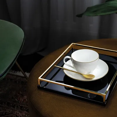 Schwarzes Tablett mit goldenen Akzenten, darauf eine weiße Tasse mit goldener Umrandung der Kollektion 'Anmut' Schwarzes Tablett mit goldenen Akzenten, darauf eine weiße Tasse mit goldener Umrandung der Kollektion 'Anmut' von Villeroy & Boch und einem Löffel.