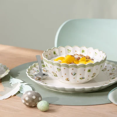 Easter Delight bowl with Easter motifs A decorative Easter Delight bowl with fruit and yogurt stands on a plate with a spoon next to a cup and saucer on a table.