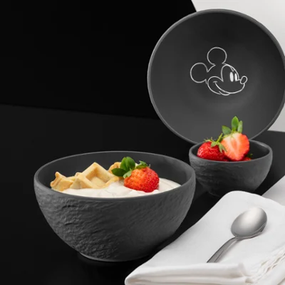 Czarna, teksturowana miska z kolekcji Manufacture Mickey, wypełniona jogurtem, truskawkami i kawałkami wafli, obok dopasowanej pokrywki z motywem Myszki Miki. Łyżka i starannie złożona serwetka leżą obok misek na czarno-białej powierzchni.