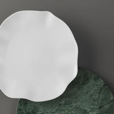 Ein weißer Pura Teller mit gezacktem Rand über einem dunkelgrünen, rundem Tablett Ein weißer Pura Teller mit gezacktem Rand wird über ein dunkelgrünes, rundes Tablett auf einer grauen Fläche gelegt.