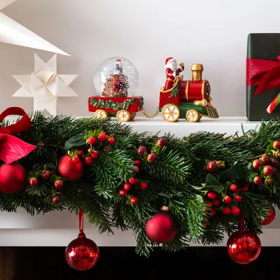 A festively decorated mantelpiece with the decorative train from the Christmas Toy collection by Villeroy & Boch. With red ornaments of a snow globe, star-shaped decorations and a green gift box decorated with a red bow and a candy cane.