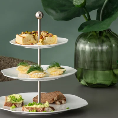Three-tiered étagère from the Pura collection with various finger foods on a table Three-tiered étagère from the Pura collection with various finger foods on a table, with green glass vases and plants in the background.