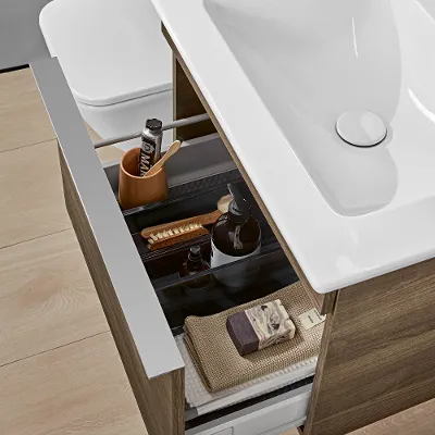 Venticello Waschbecken mit geöffneter Badezimmerschublade Unter einem weißen Venticello Waschbecken ist eine Badezimmerschublade geöffnet und gibt den Blick auf geordnete Toilettenartikel frei, darunter eine Seifenschale, eine Zahnbürste, einen Rasierer und Handtücher
