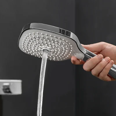 Une main tient une douchette d'où s'écoule de l'eau, devant un fond de salle de bains sombre.
