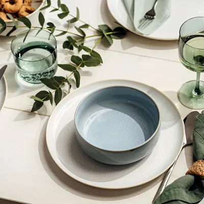 Graziosa tavola apparecchiata con una ciotola azzurra di Crafted di Villeroy & Boch su un piatto bianco, bicchieri verdi, rami di eucalipto e tovaglioli di stoffa verde con anelli. Altri piatti, posate e cibo sono parzialmente visibili.