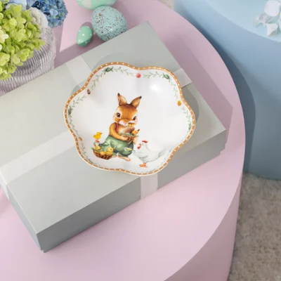A decorative Annual Easter Edition 2026 plate A gift box with a ribbon sits on a pink table, topped with a decorative Annual Easter Edition 2026 plate featuring a rabbit and flowers.