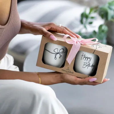 Person holding gift box with pink bow, containing two 'Happy Birthday' statement mugs.