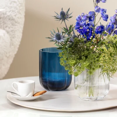 A small white For me cup with a biscuit on a saucer, next to a blue Fleur vase and a transparent Fleur vase A small white For me cup with a biscuit on a saucer, next to a blue Fleur vase and a transparent Fleur vase with blue and green flowers on a round marble surface.