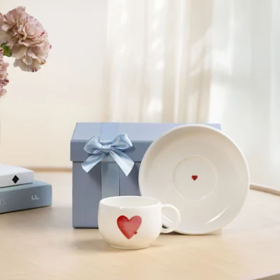 Filiżanka z podstawką With Love jako pomysł na prezent Biała filiżanka With Love z czerwonym motywem serca, dopasowana podstawka i niebieskie pudełko prezentowe z kokardką na drewnianym stole przy oknie.