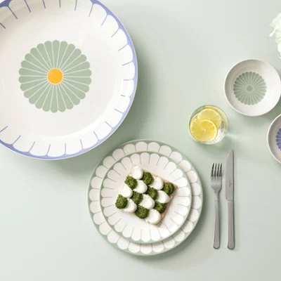A laid table with decorative plates and bowls, a slice of bread with vegetables on a plate, a fork and knife next to it and white flowers on the right.