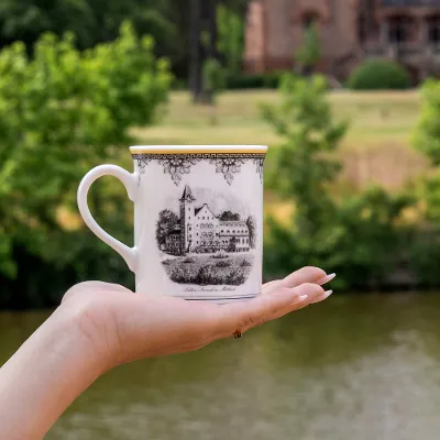 One hand holds a decorative Audun cup with an illustration of a building in front of a river and trees.