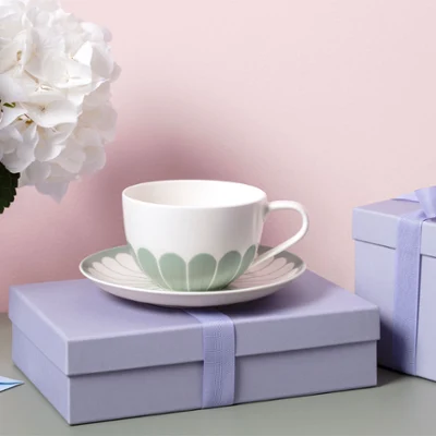 A Fleur teacup and saucer stands on a lavender-coloured gift box next to a matching box and a vase of flowers.