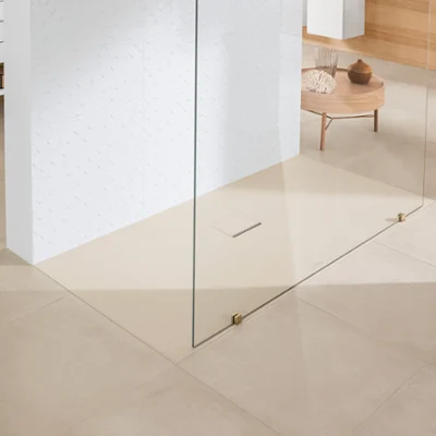Une douche à l’italienne Squaro Infinity dans une salle de bains avec un carrelage de sol beige Une salle de bains minimaliste avec des carreaux de sol beiges, une douche à l’italienne séparée par une plaque en verre, un mur carrelé blanc et une petite table en bois avec des objets décoratifs en arrière-plan.