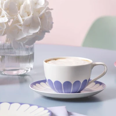 The blue Fleur cup of coffee with foam stands on a patterned saucer next to a vase of white flowers on a pastel-coloured table.
