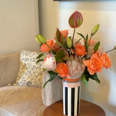 Memphis vase with flowers on side table Colorful floral arrangement of orange roses, lilies, and green buds in a striped vase on a wooden table beside a cozy chair, warm interior.