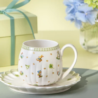 Easter Delight teacup on a Easter Delight saucer Easter Delight teacup on a Easter Delight saucer with a gift box and a vase of blue flowers in the background.